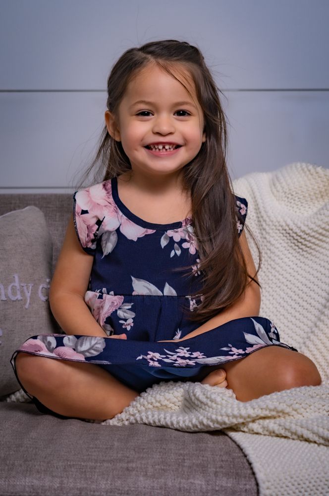 A child in a floral navy dress sits on a grey couch with a white blanket.