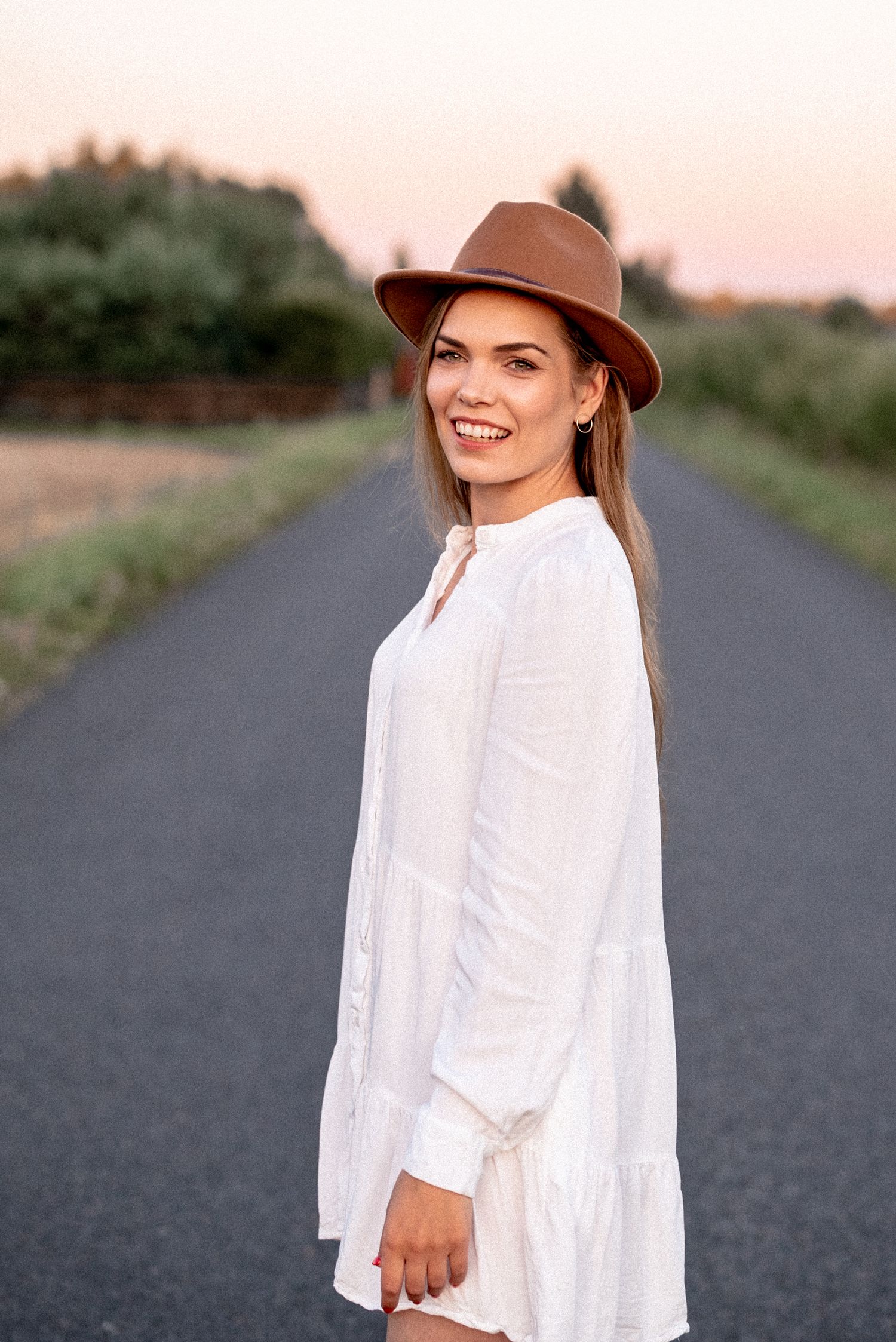 A woman in a white dress and brown hat poses on a country road at sunset.