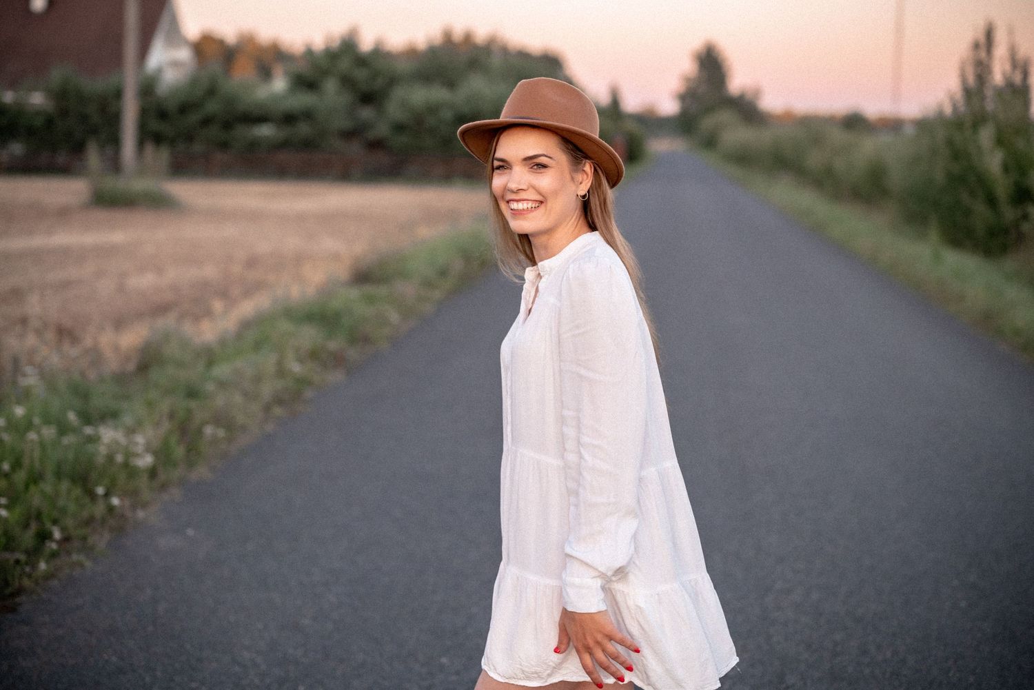 Someone wearing a casual white dress and brown hat stands on an asphalt path during sunset.