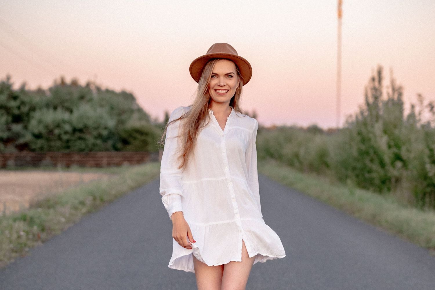 Someone wearing a white summer dress and hat smiles while standing on a country road.