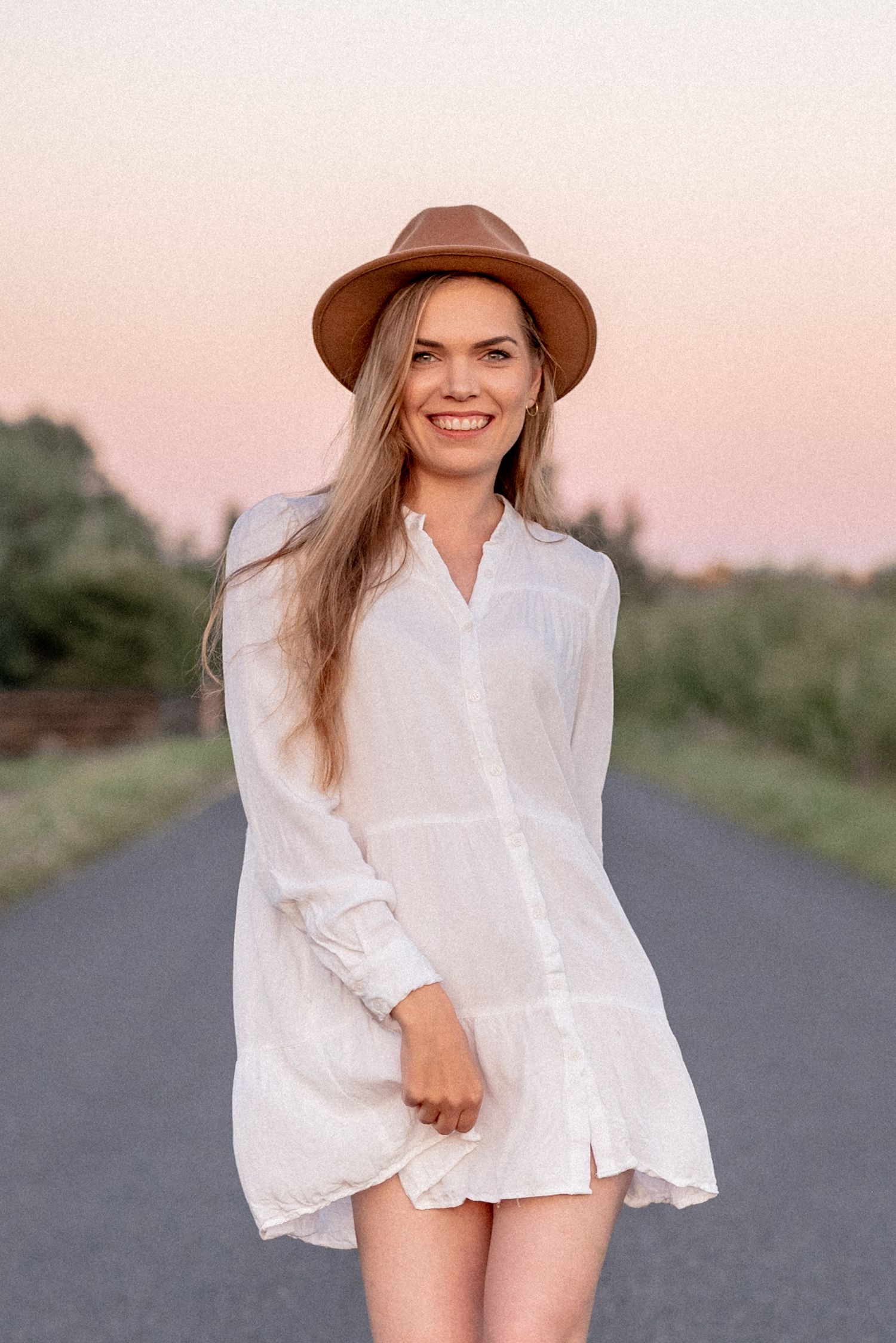 A portrait of a person in a white shirt dress and brown fedora on a rural road at sunset.