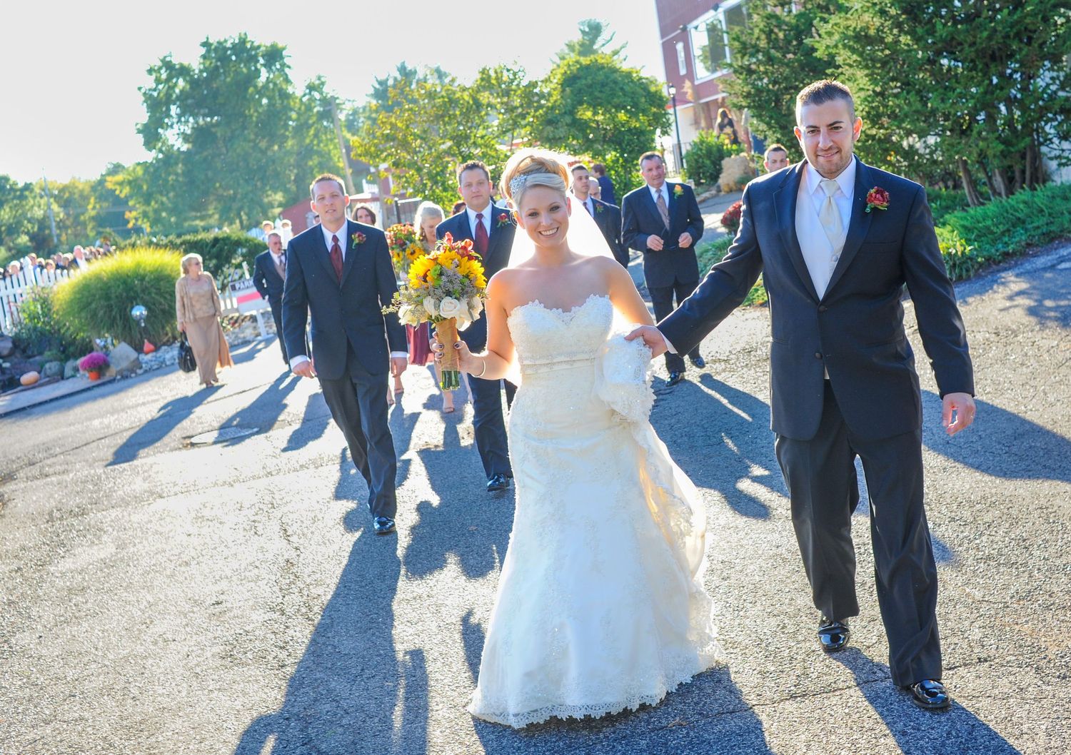 Wedding party walks down sunny path with bride in white dress and yellow flowers.