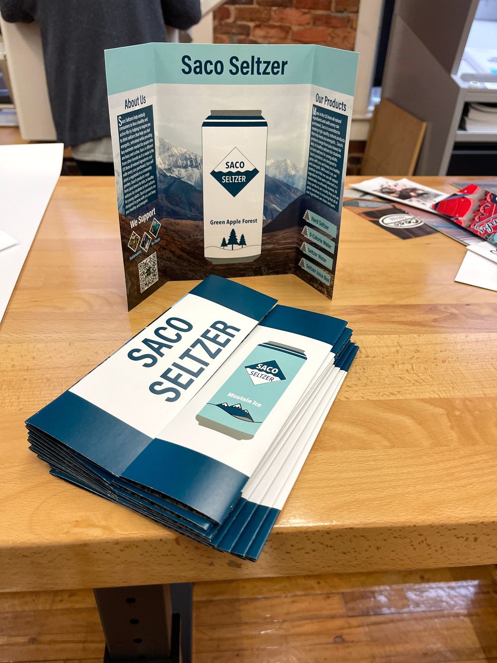 Marketing materials for Saco Seltzer displayed on a wooden table showing branded brochures and promotional items.
