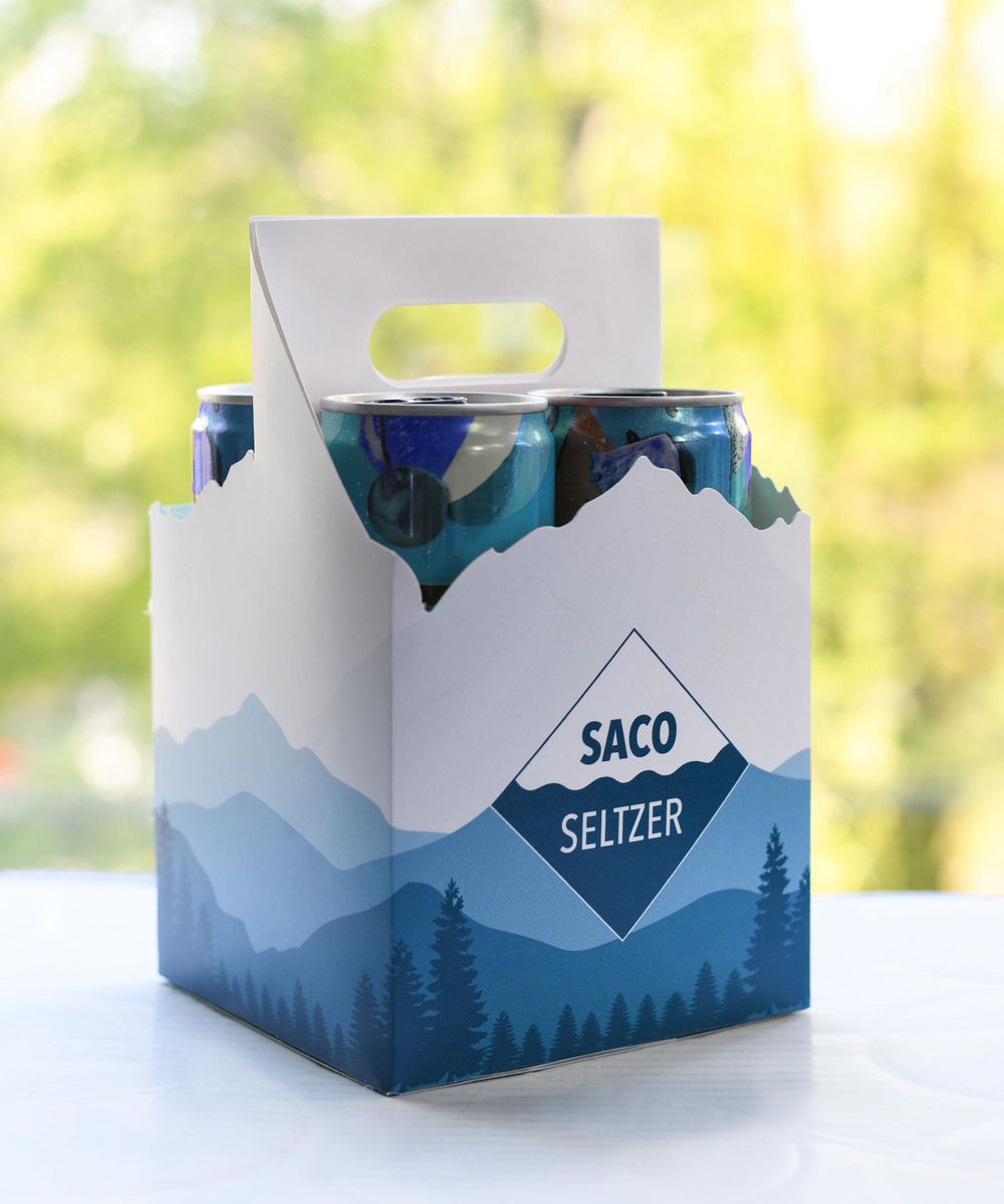 White cardboard carrier package for Saco Seltzer featuring mountain landscape design in blue tones.