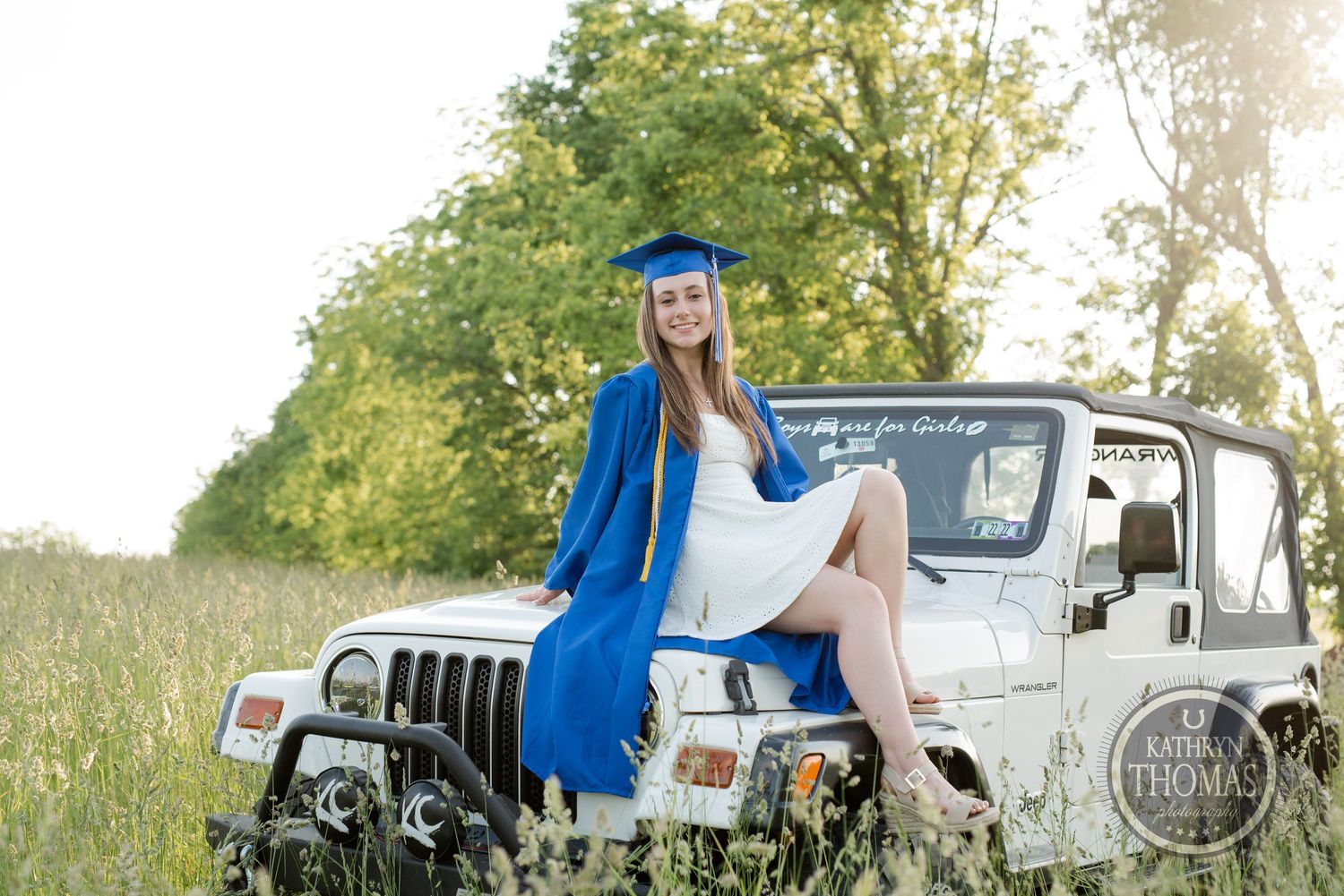 Graduate in blue cap and gown poses on vintage Jeep in sunny field with trees in background.