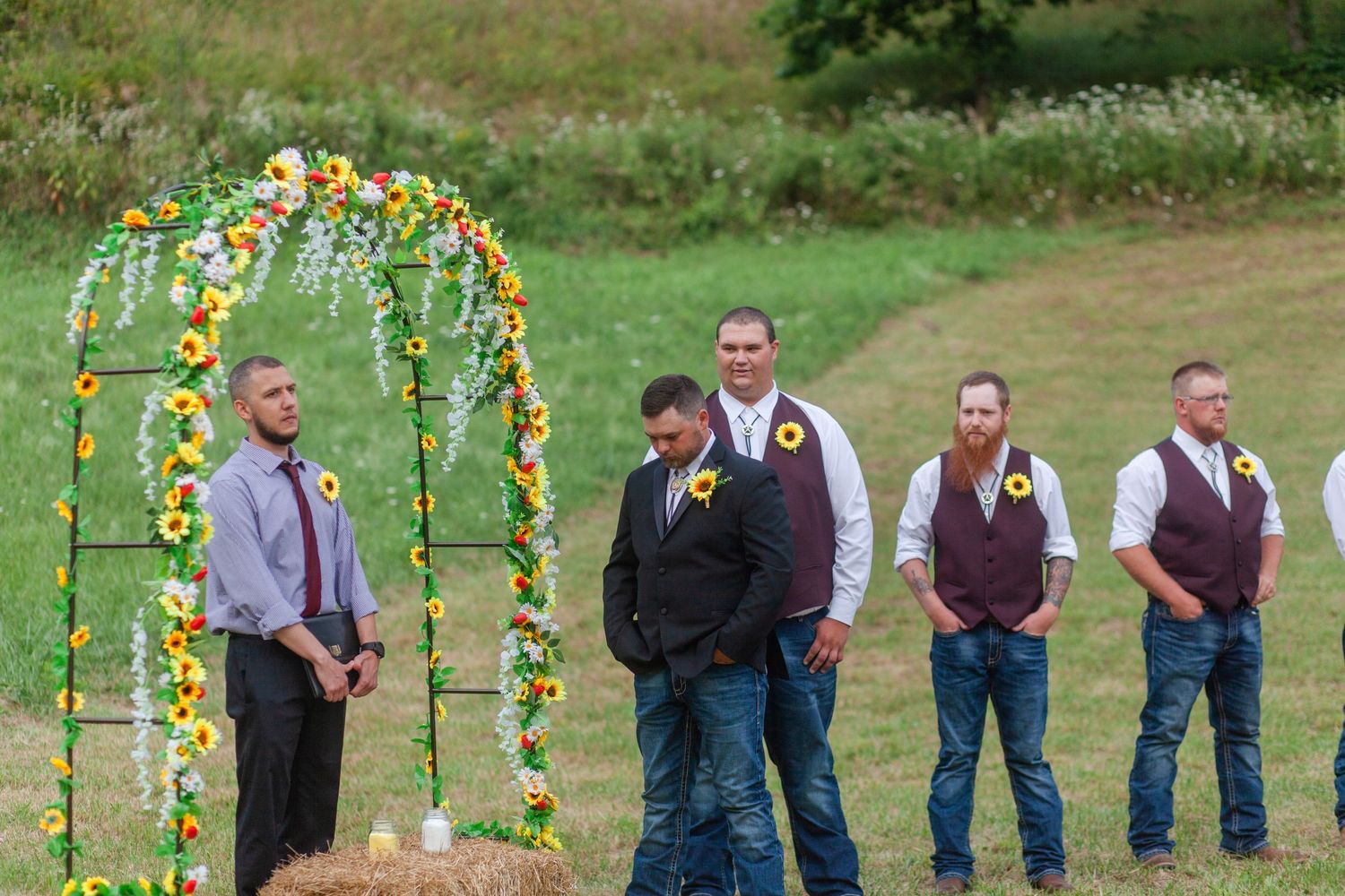 Wedding party members gather under a sunflower-decorated arch in an outdoor field setting.