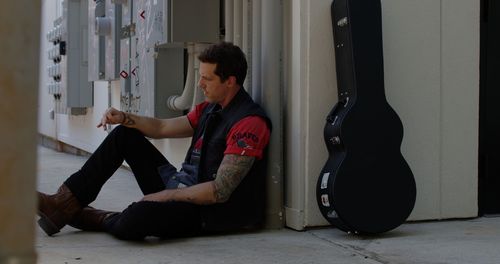 Person sits against wall near guitar case in hallway corner.
