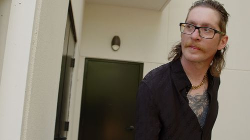 Individual with glasses and tattoos in dark clothing standing in hallway.