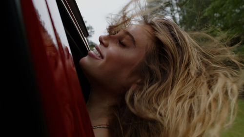 Person with blonde hair leaning against a red vehicle.