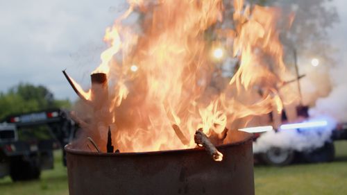 Large flames rise from a metal fire pit at an outdoor event.