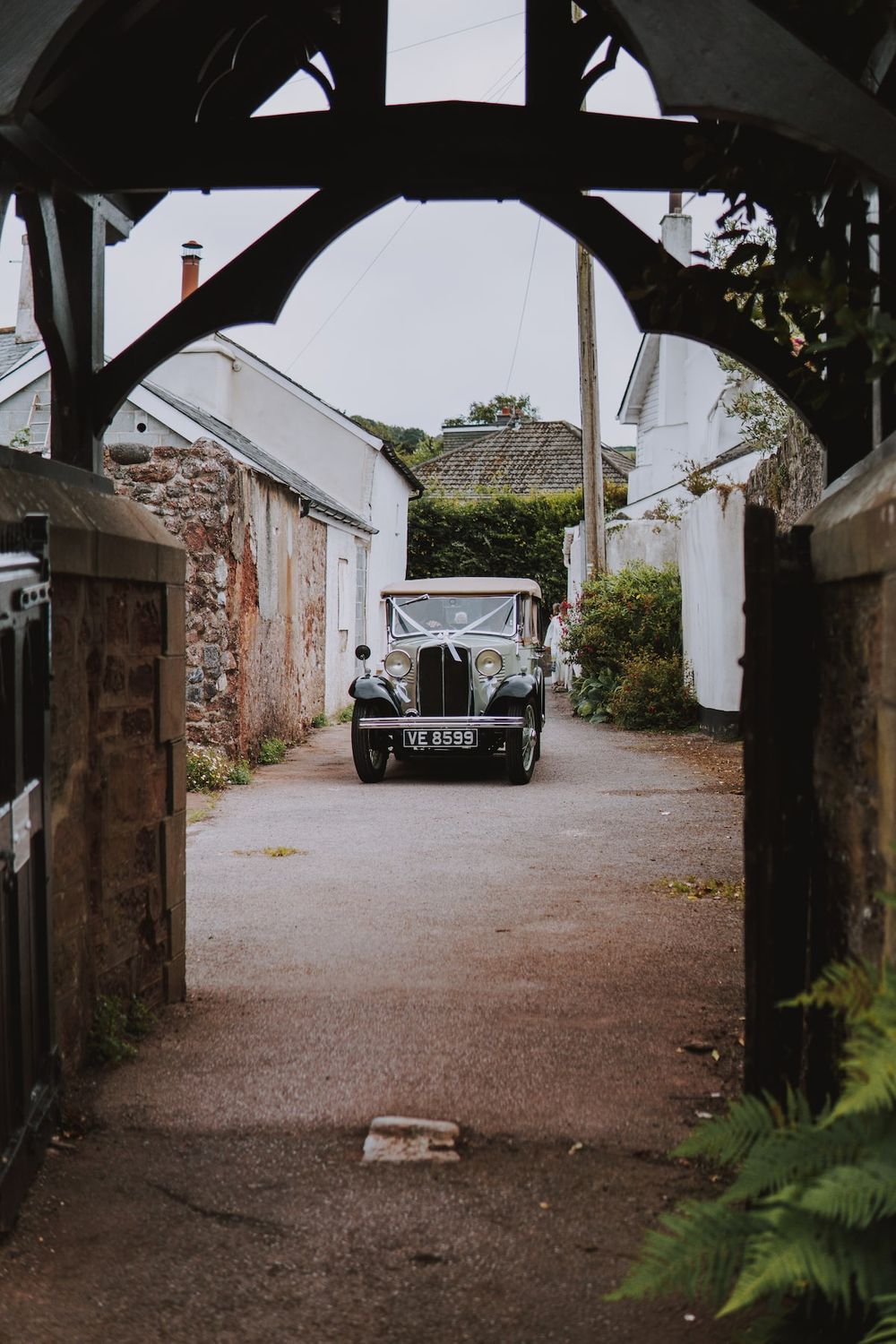 Vintage car viewed through wooden archway in rustic stone courtyard.