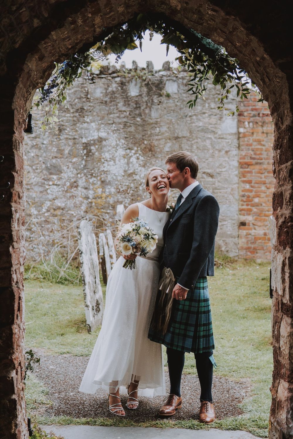 A romantic wedding moment beneath stone archways showing a couple in traditional Scottish attire with a white dress and kilt.