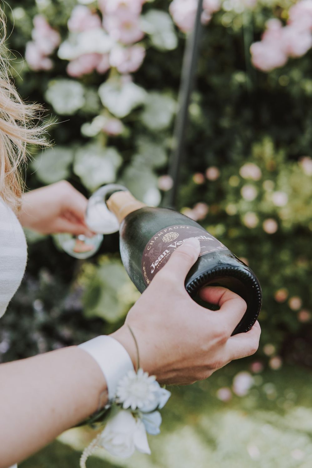 Hands opening a champagne bottle in a garden with pink flowers in the background.