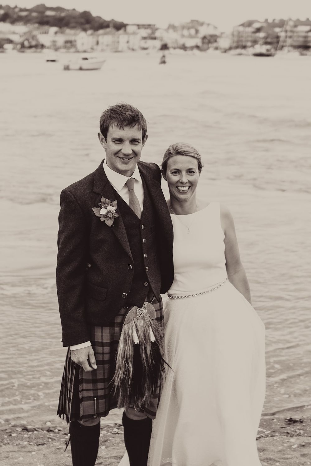 Black and white portrait of newlyweds in Scottish wedding attire on a beach.