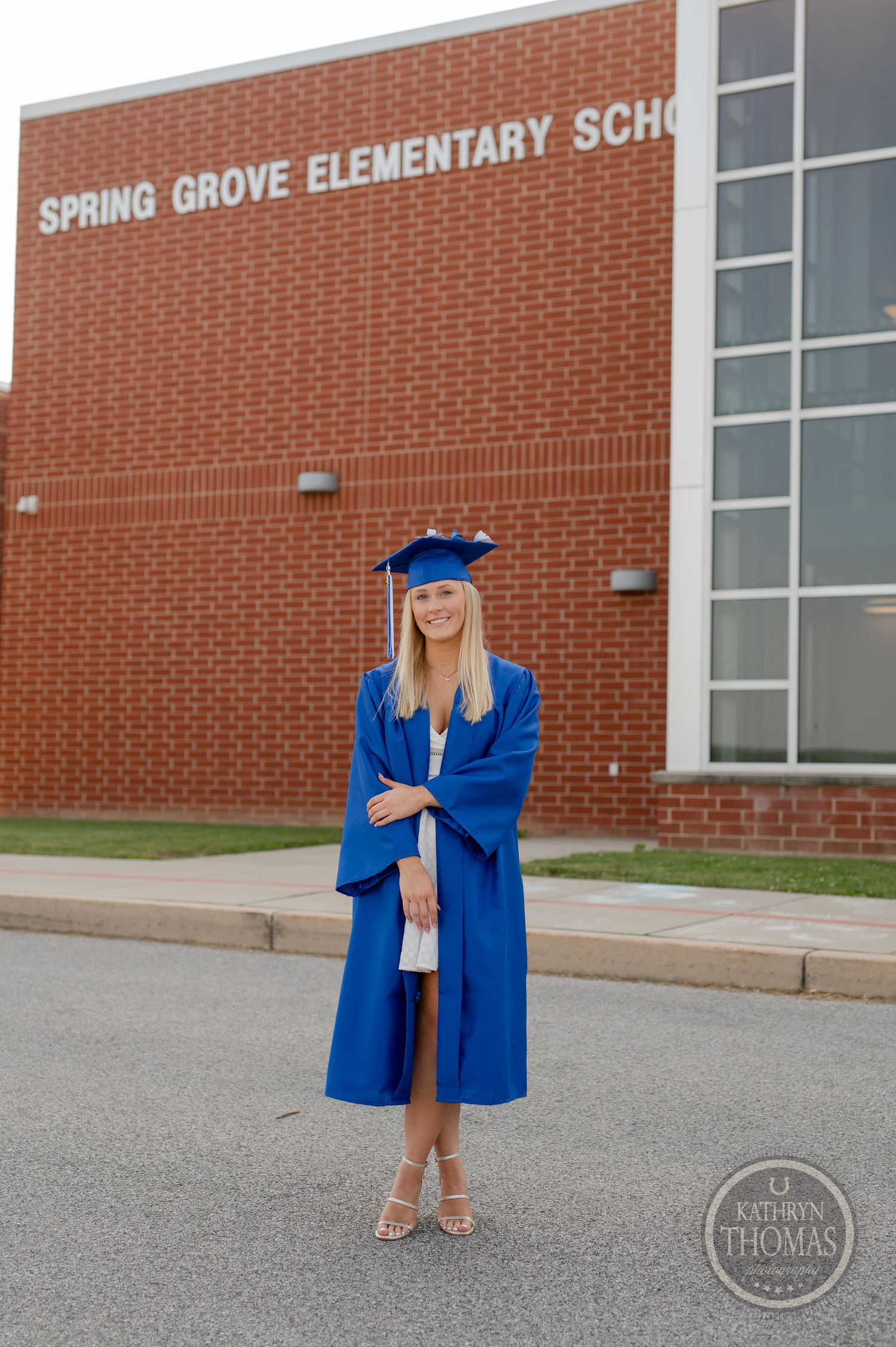 Graduate in blue cap and gown stands in front of brick elementary school building.