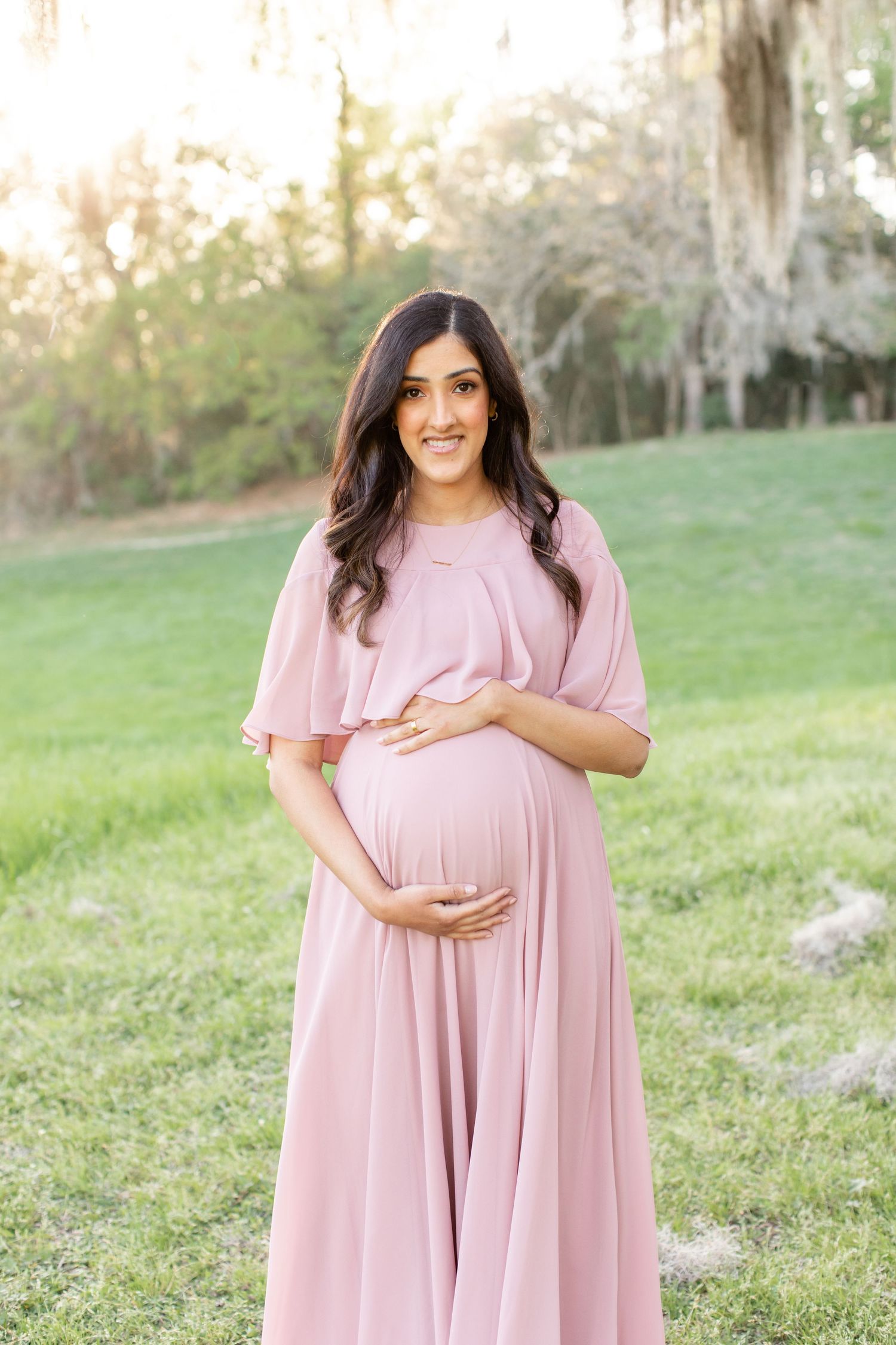Expectant mother in flowing pink maxi dress poses in grassy field with Spanish moss trees at sunset.