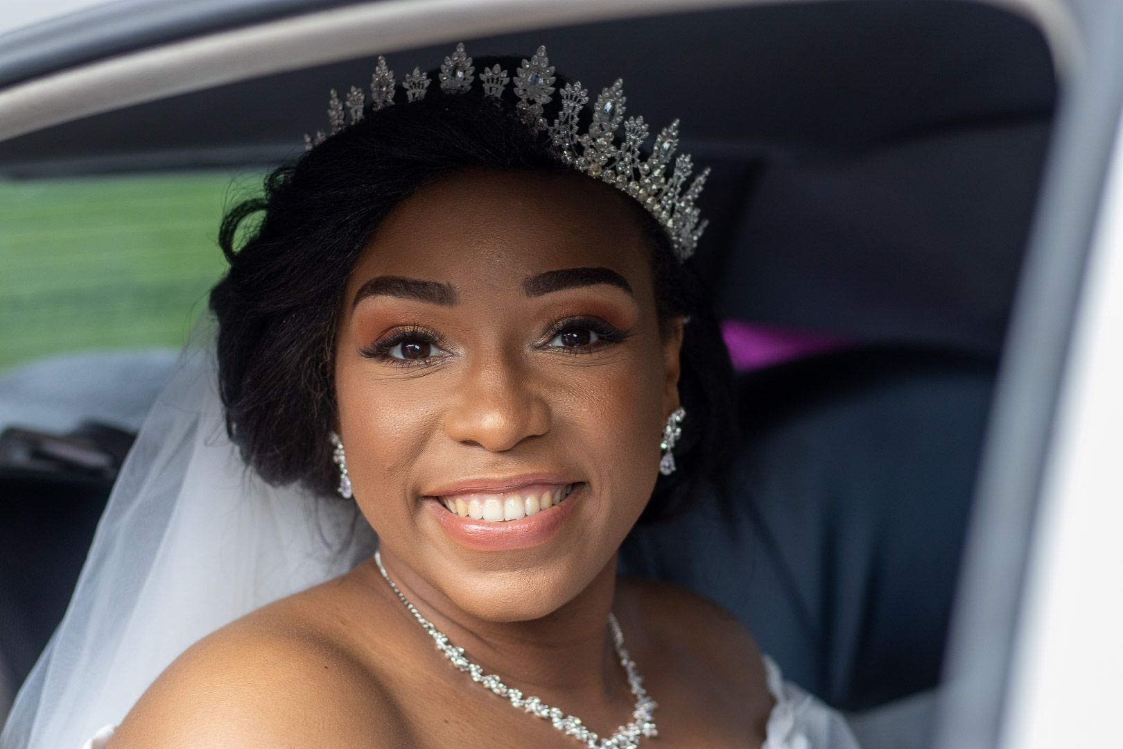 Beautiful bride wearing a sparkling tiara and necklace smiles joyfully from inside a wedding car.