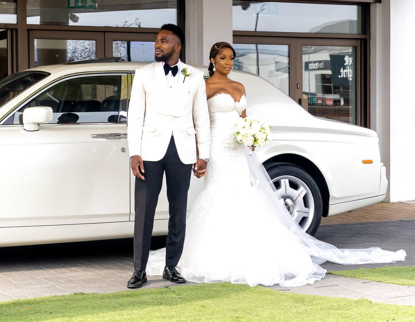 Newlyweds pose beside white luxury car, groom in white jacket and bride in flowing gown.