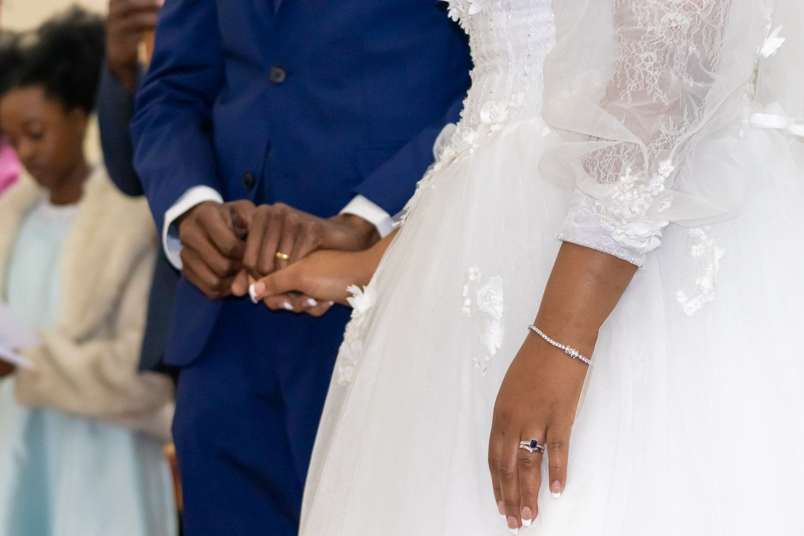 Close-up of couple holding hands during wedding ceremony, showing white dress and blue suit details.