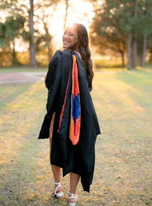 Graduate in black gown walks through autumn park during sunset portrait session.