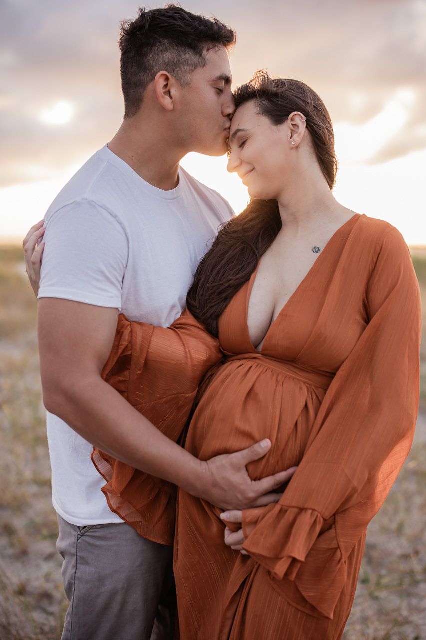 Maternity photos of couple in rust dress and white shirt during golden hour.