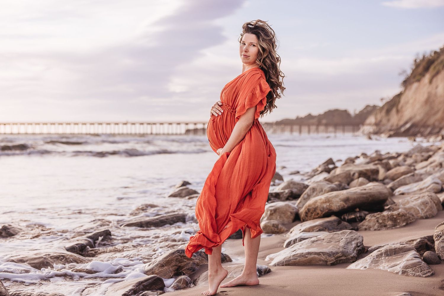 Coral maternity dress flows in ocean breeze against rocky beach landscape at sunset.