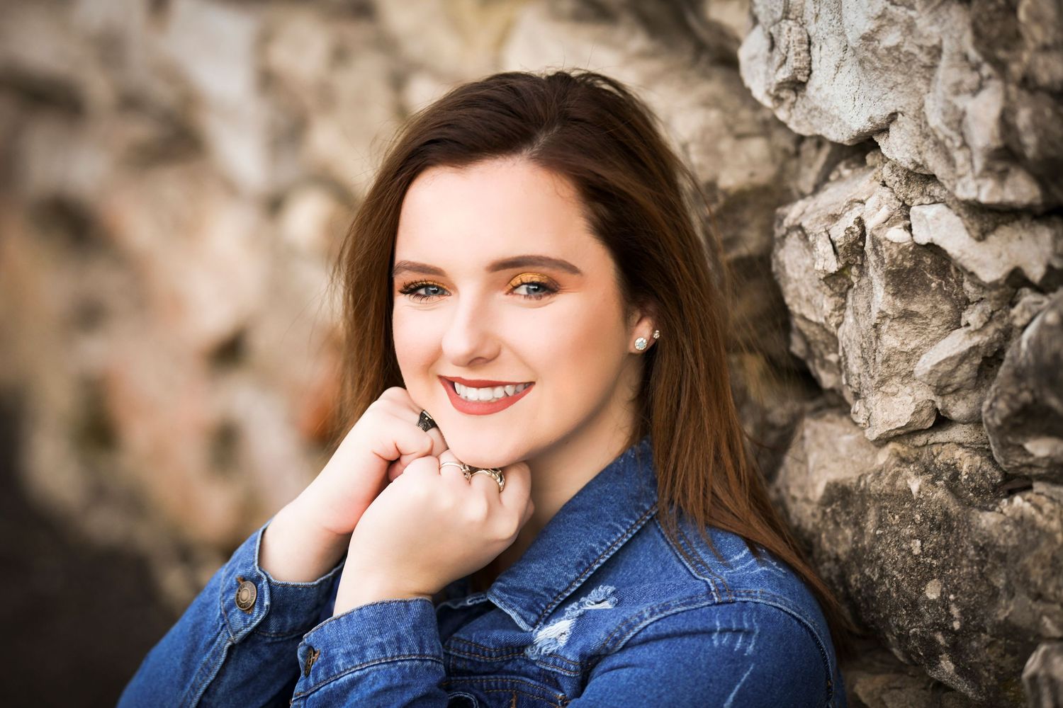 Smiling young woman in denim jacket leaning against rustic stone wall portrait.