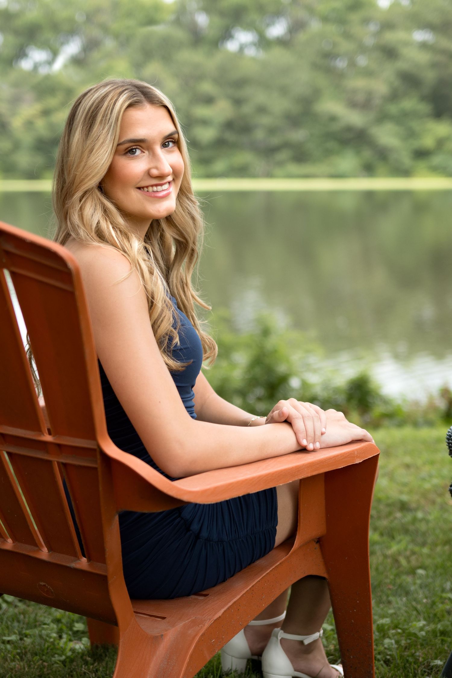 A person sits on a wooden bench near a peaceful lake in a navy dress.