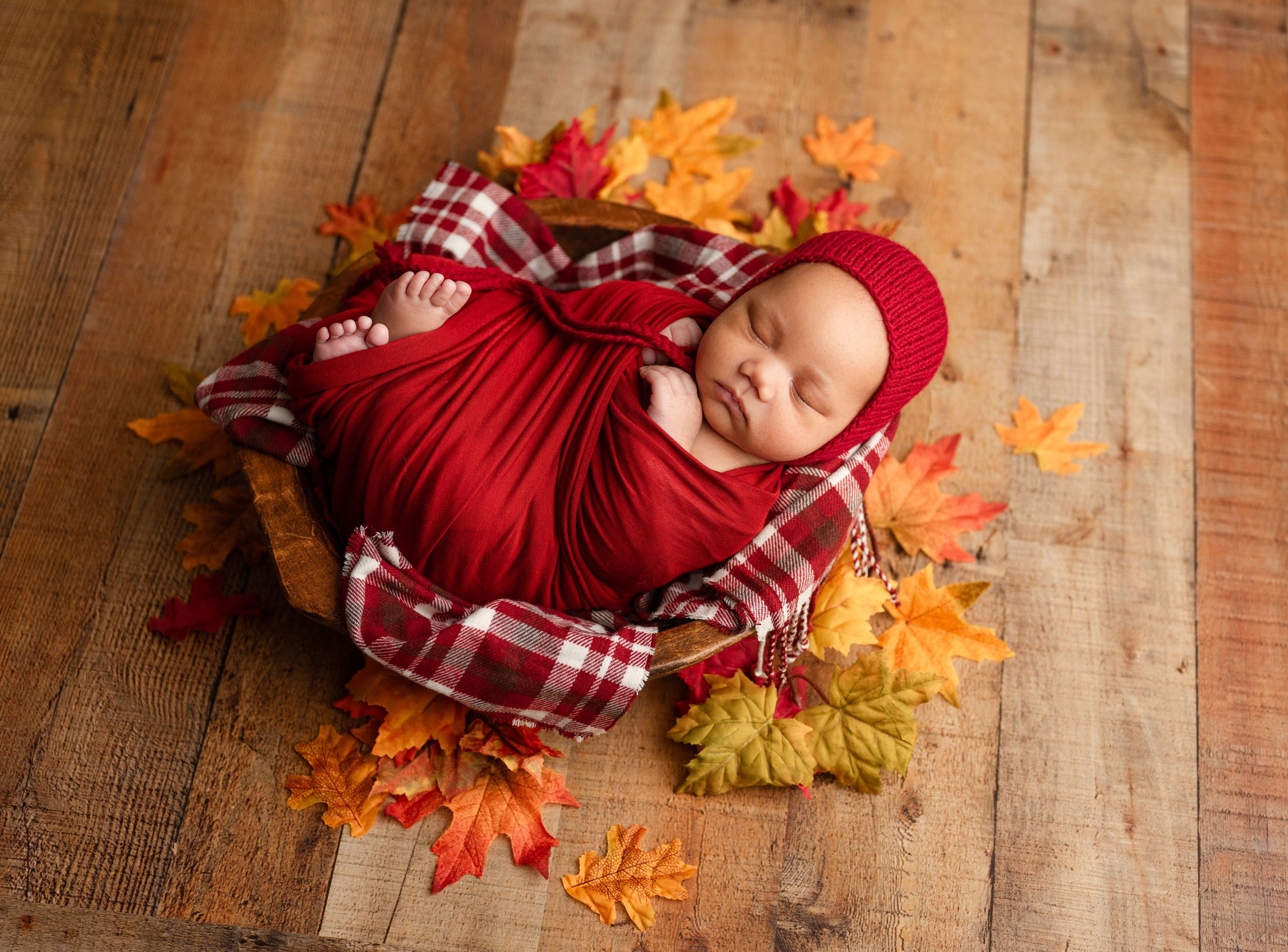 Newborn baby wrapped in red swaddle with knit hat, sleeping in bowl surrounded by colorful autumn leaves on wood floor.
