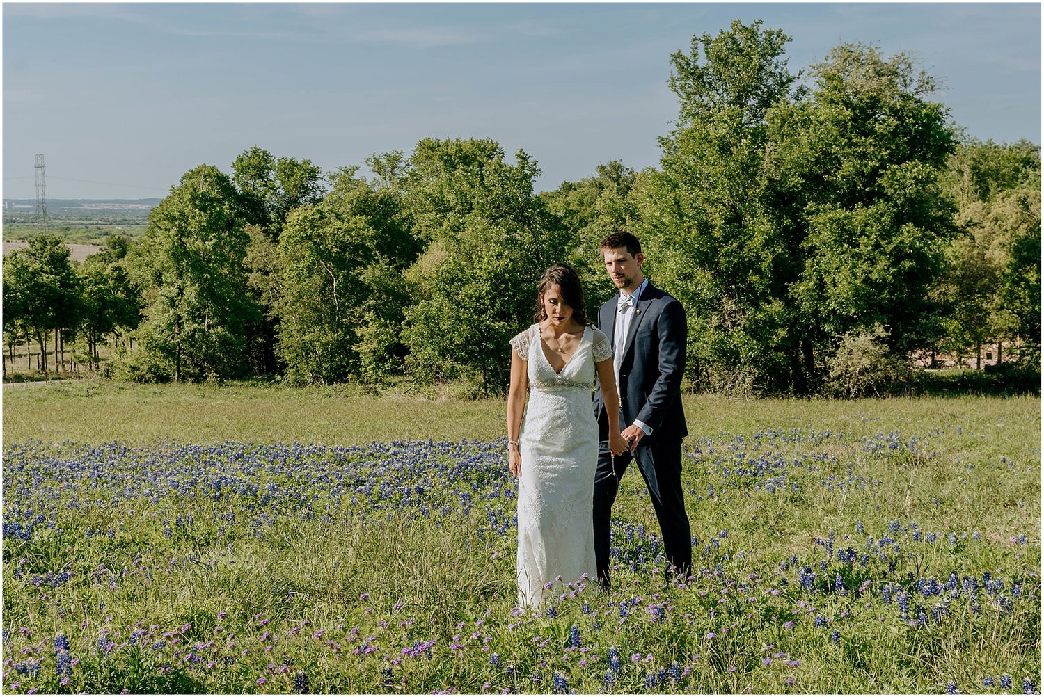 A couple stands together in a field of bluebonnets with green trees in the background on a sunny day.