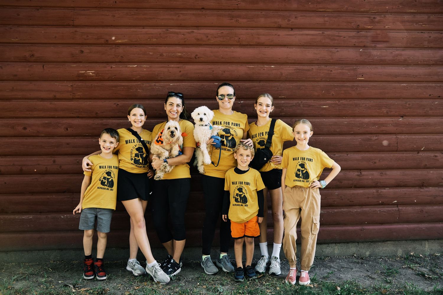 Group wearing matching yellow t-shirts poses against rustic brown wooden wall background.