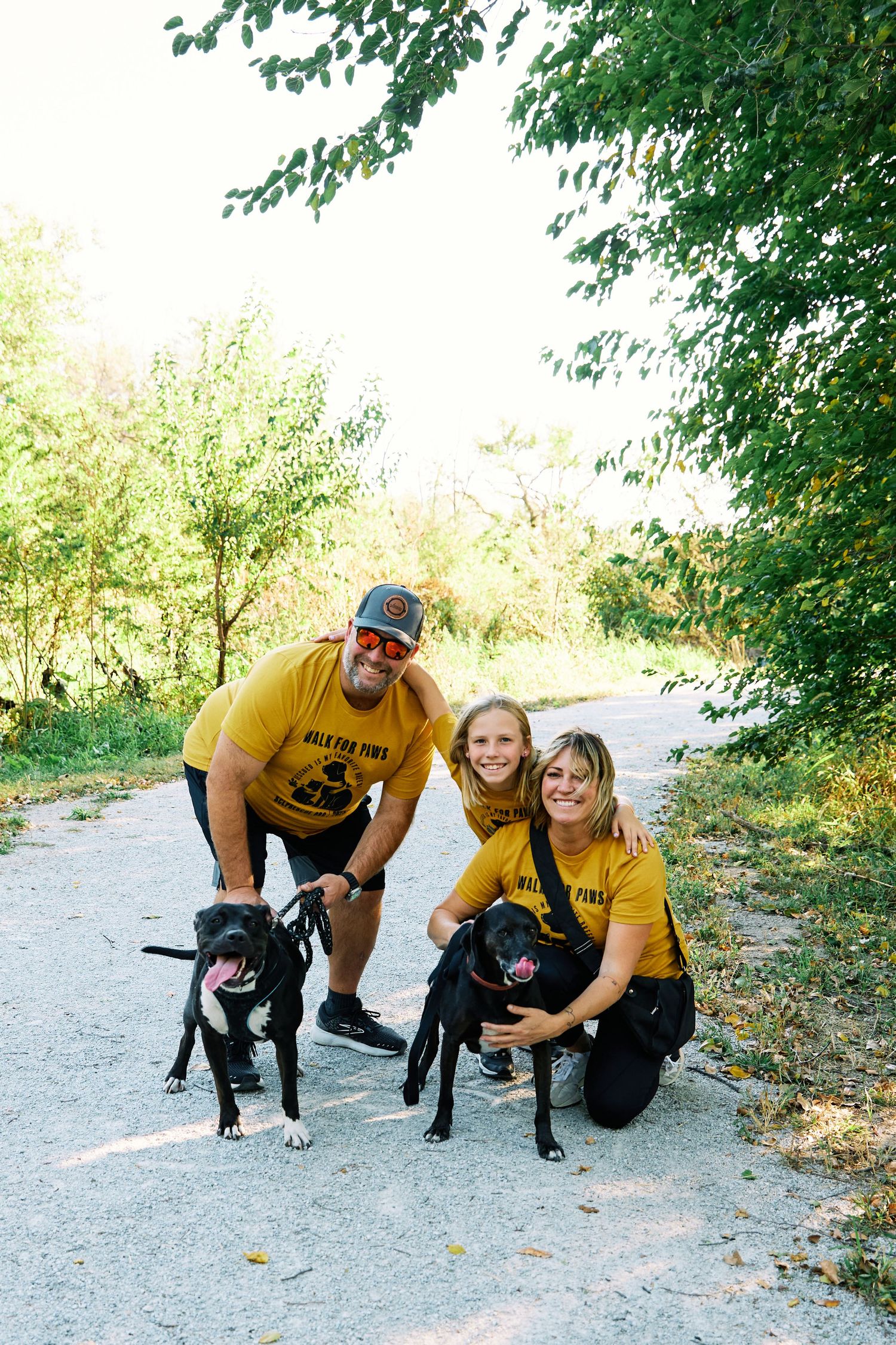 People in yellow shirts pose with black dogs on a dirt path under trees.