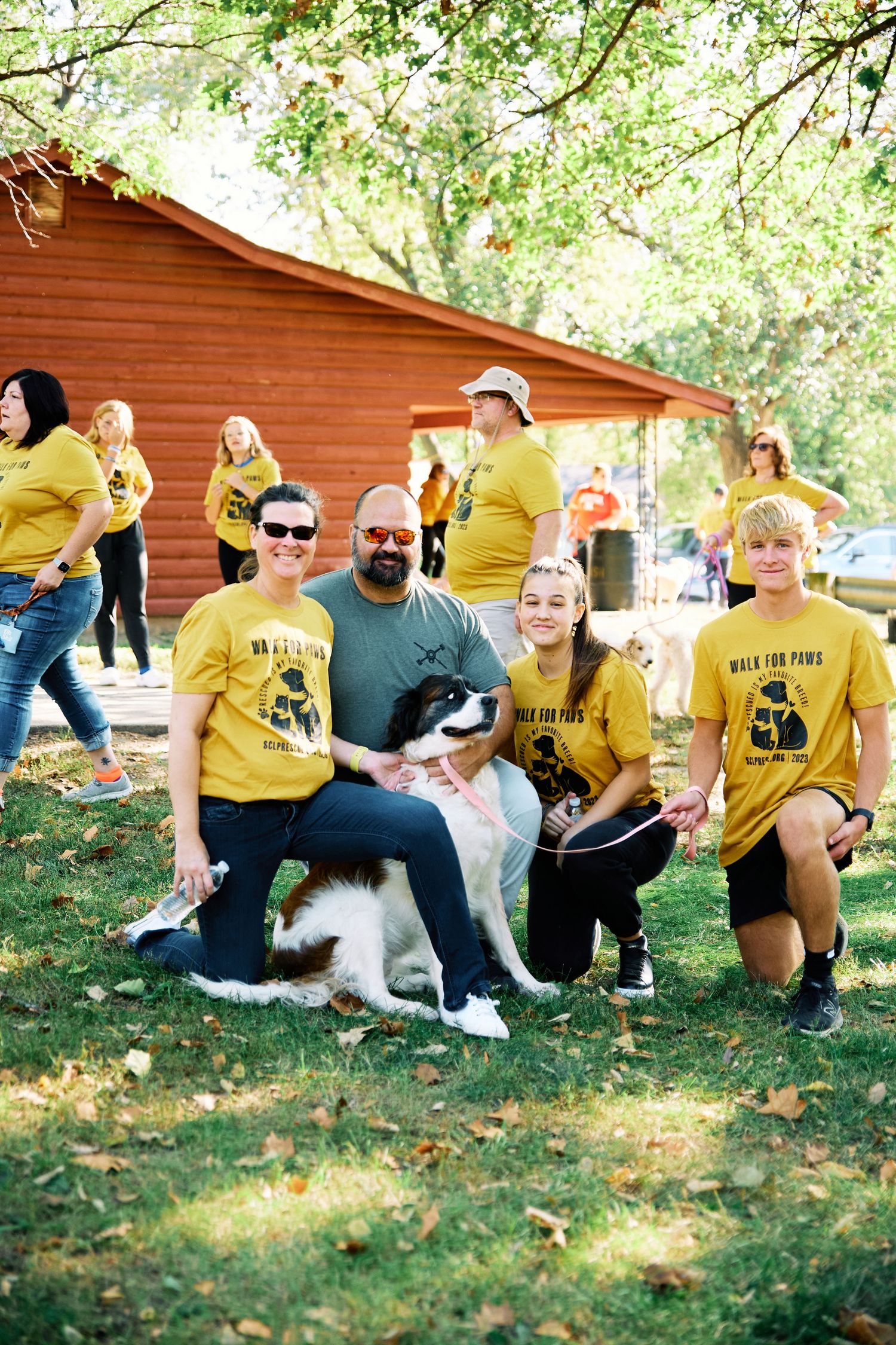 Group wearing matching yellow shirts poses with a black and white dog at an outdoor event near a red barn.