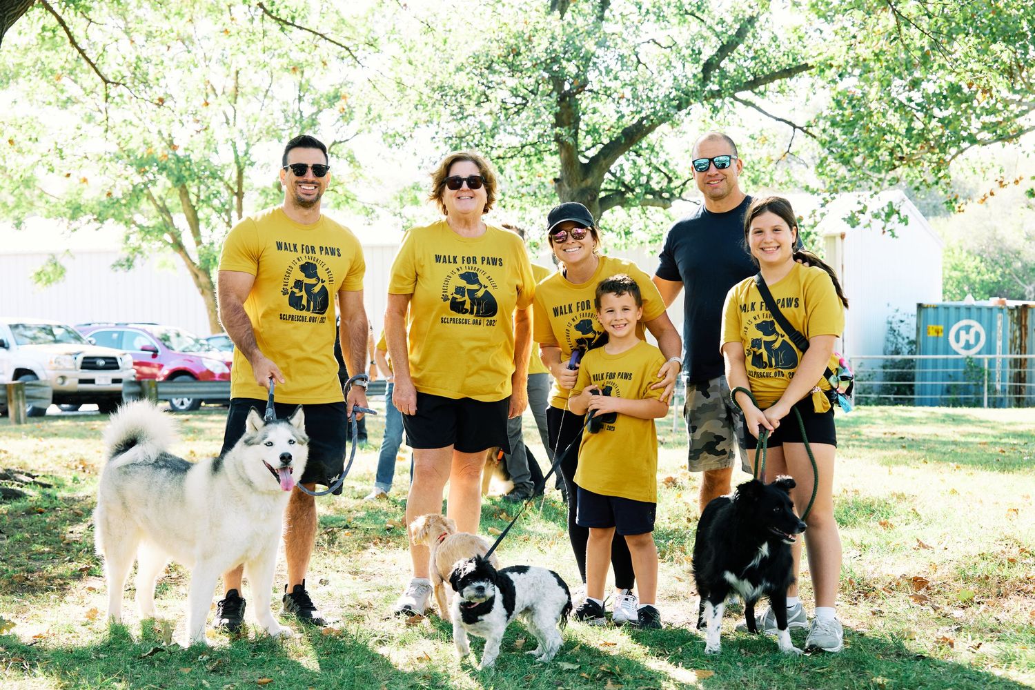 Group wearing matching yellow shirts poses with their various dogs at a park event.
