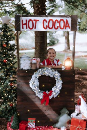 Christmas hot cocoa stand setup with white wreath and red bow decorations against wooden backdrop.