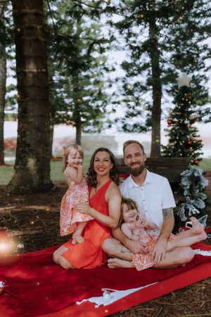 Family enjoying a holiday photoshoot on a red blanket under pine trees.