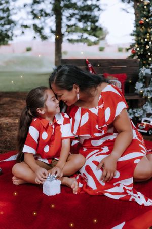 Mother and daughter in matching red and white outfits sharing a tender moment by Christmas tree.