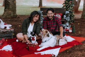 A couple and their pets pose with Christmas decorations on a red blanket outdoors.