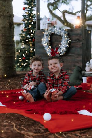 Children in plaid shirts lay on a red blanket near a decorated Christmas tree and holiday decor.