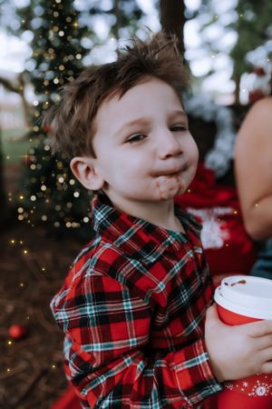 A child in a red plaid shirt smiles while holding a holiday drink cup near a Christmas tree.