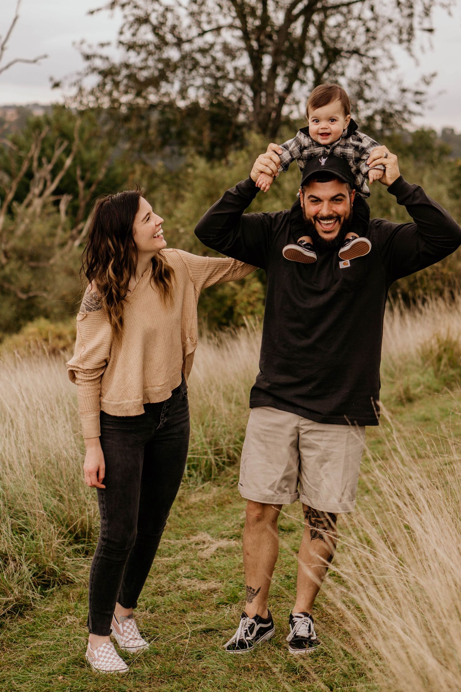 Happy family outdoors, father carrying toddler on shoulders while mother smiles in grassy field.