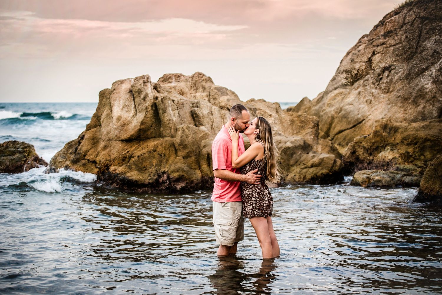 A romantic couple embraces while standing in shallow ocean water surrounded by rocky coastal formations at sunset.