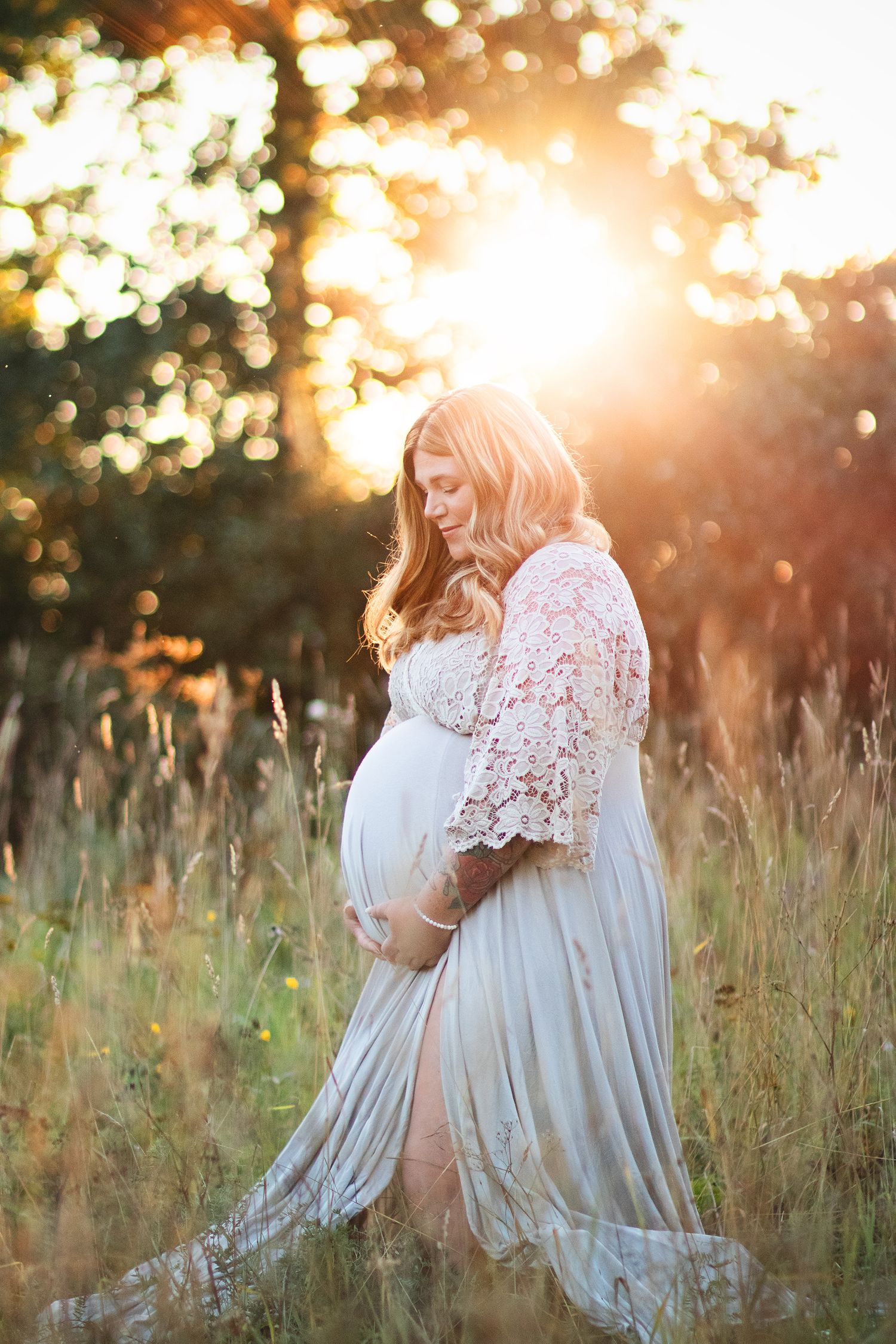 Sunlit maternity portrait features white lace dress in golden field.