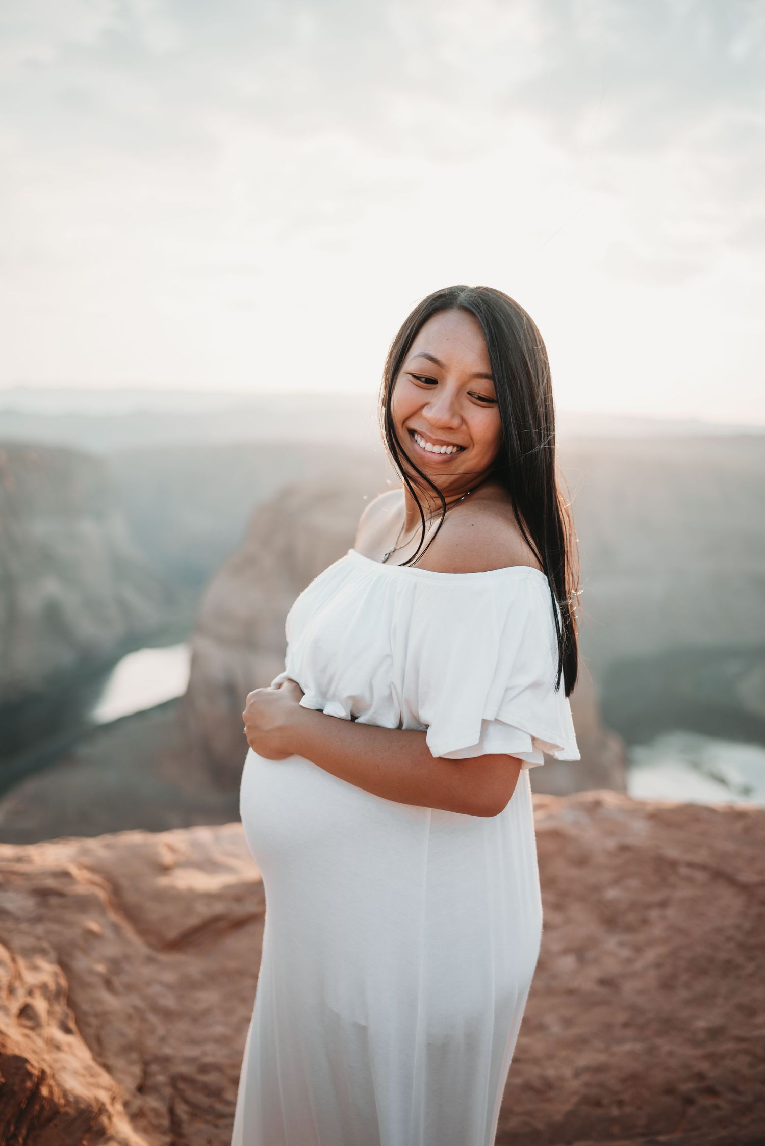 A woman in a flowing white dress stands on red rocks at sunset overlooking a canyon vista.