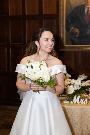 Radiant bride smiles while holding a beautiful white rose bouquet in an elegant wood-paneled reception hall.