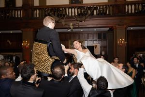 Bride and groom are lifted by guests in chairs during a lively wedding reception celebration.