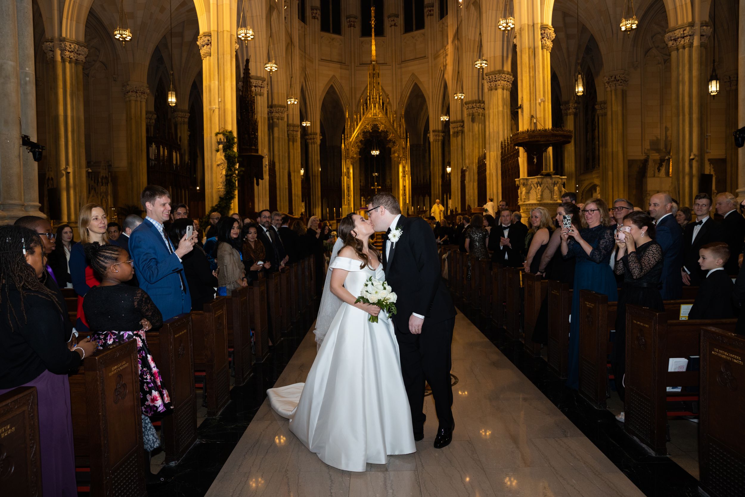 Newlyweds share a romantic kiss in the aisle of a grand cathedral as guests applaud.