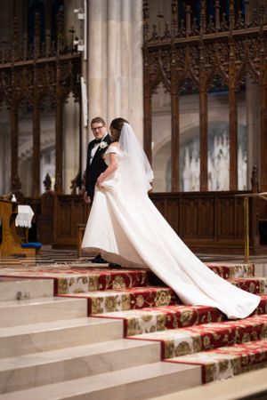 Bride in a flowing white gown with long train walks up red-carpeted altar steps with groom.