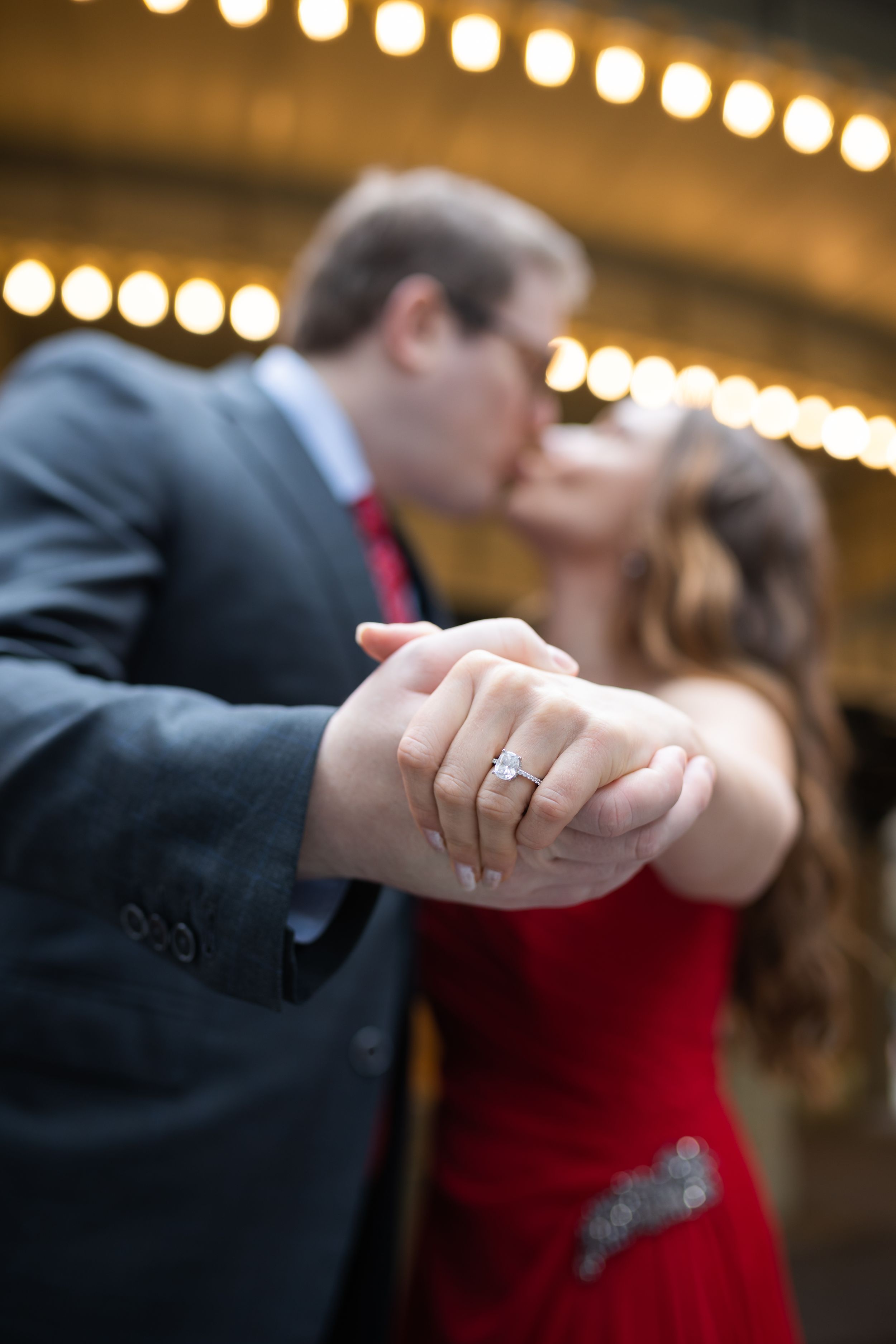 Couple kisses while showcasing a sparkling diamond engagement ring under warm marquee lights.