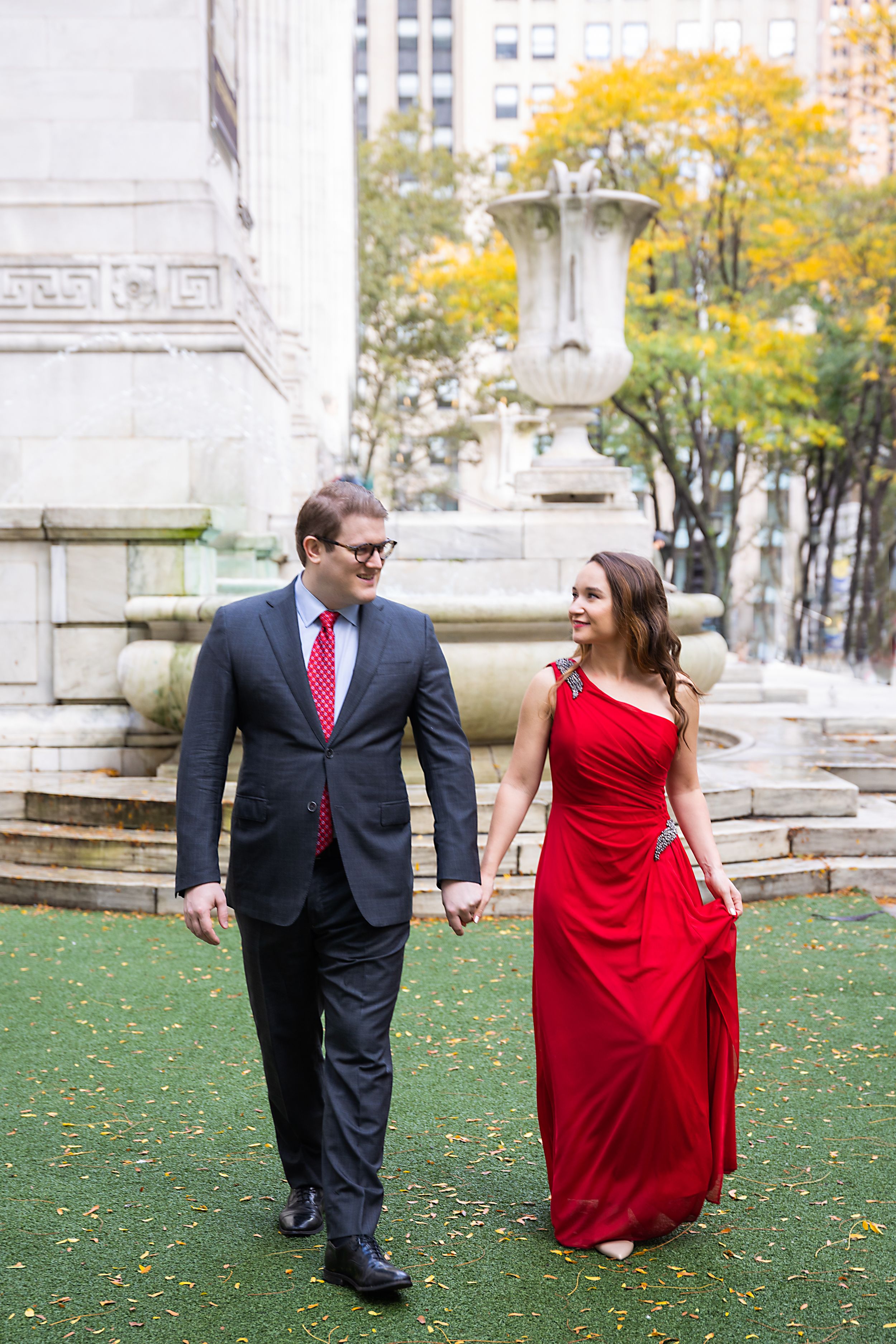 Couple holding hands walking in Bryant Park, woman in red gown and man in gray suit with red tie.